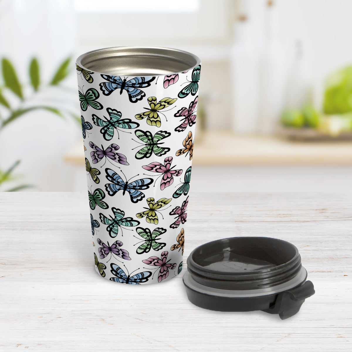 Pretty Butterfly Pattern Travel Mug (15oz) at Amy's Coffee Mugs. A stainless steel travel mug designed with pretty and colorful butterflies in a pattern that wraps around the mug. This travel mug is perfect for people who love butterflies and colorful nature designs. Photo shows the tapered mug with the lid off, beside it on the table.