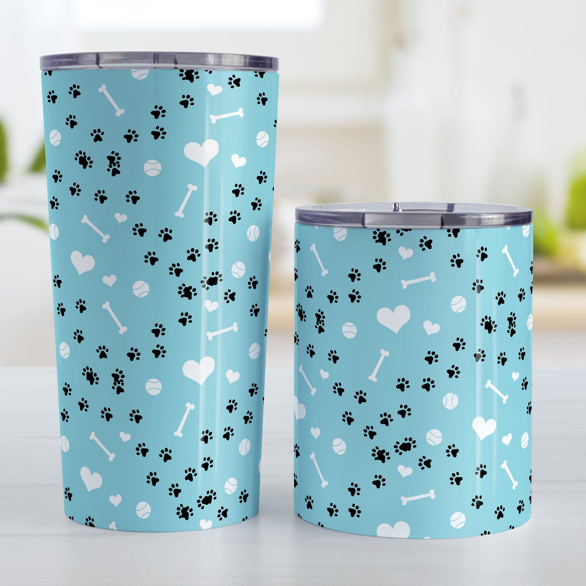 Puppy Run Blue Dog Tumbler Cup (20oz and 10oz) at Amy's Coffee Mugs. Stainless steel insulated tumbler cups designed with a pattern of chaotic tracks of puppy paw prints running around the cup with white dog bones, playing balls, and hearts over a light blue background that wraps around the cups. These cups are perfect for people who love puppy dogs, cute paw print designs, and the color blue. Photo shows both sized cups next to each other. 
