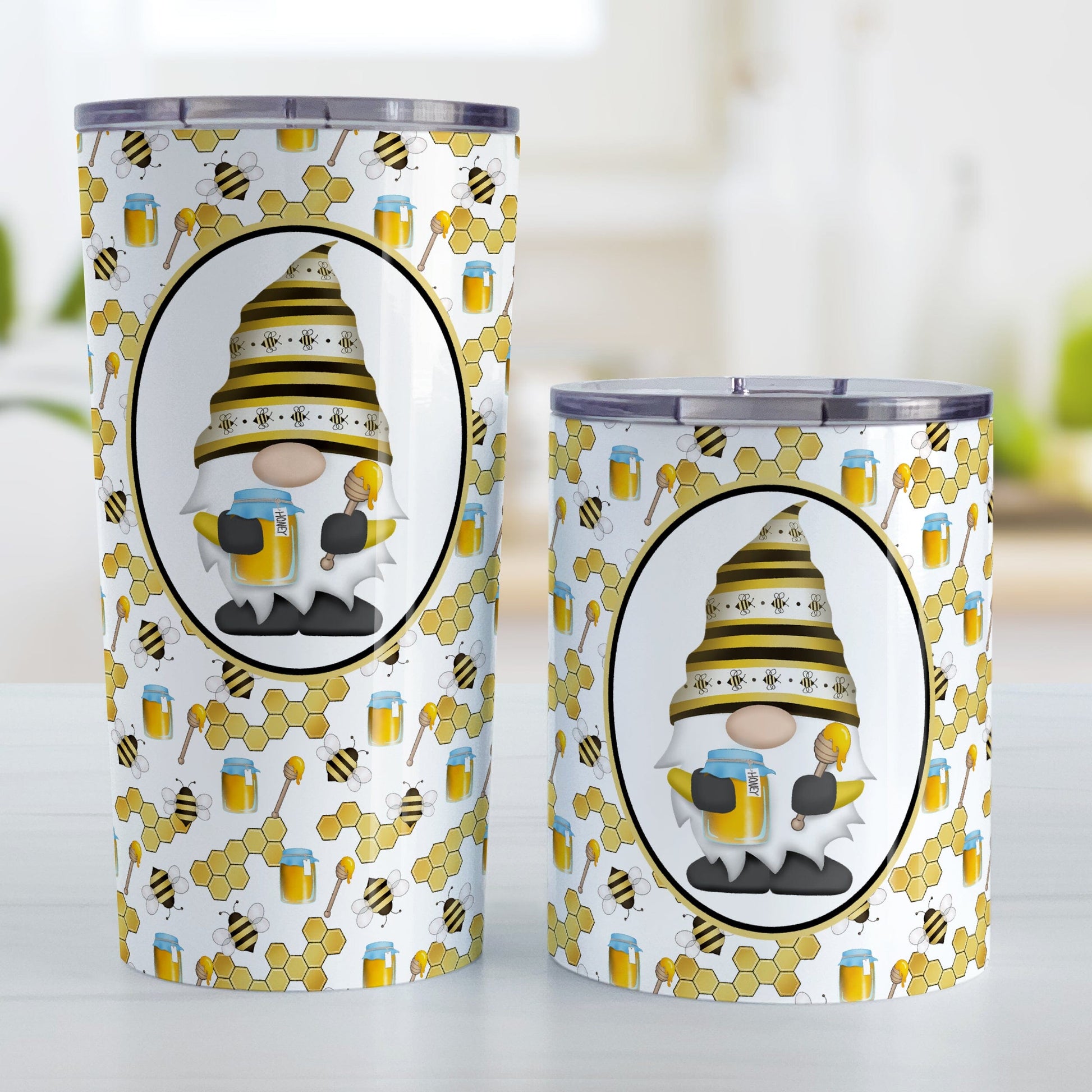 Yellow Gnome Honey Jar Bee Tumbler Cup (20oz or 10oz) at Amy's Coffee Mugs. Stainless steel tumbler cups designed with an adorable gnome wearing a yellow bee-themed hat and holding a honey jar and honey dipper in a white oval over a pattern background that wrap around the cups with bees, honey jars, honey dippers, and honeycomb. Photo shows both sized cups on a table next to each other.